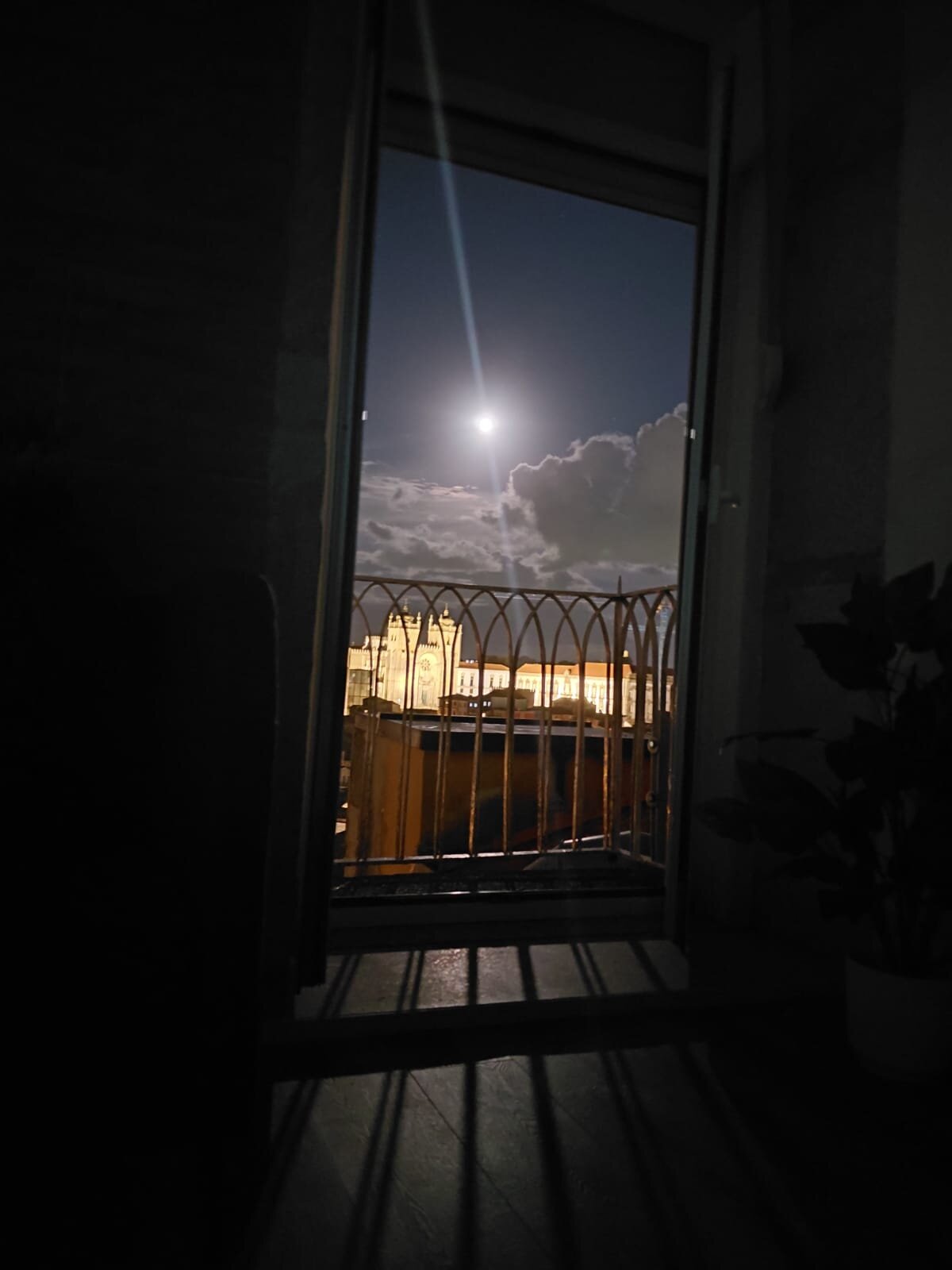 View from the balcony framed by the window and iron railing, with the illuminated Sé Cathedral and full moon in the background